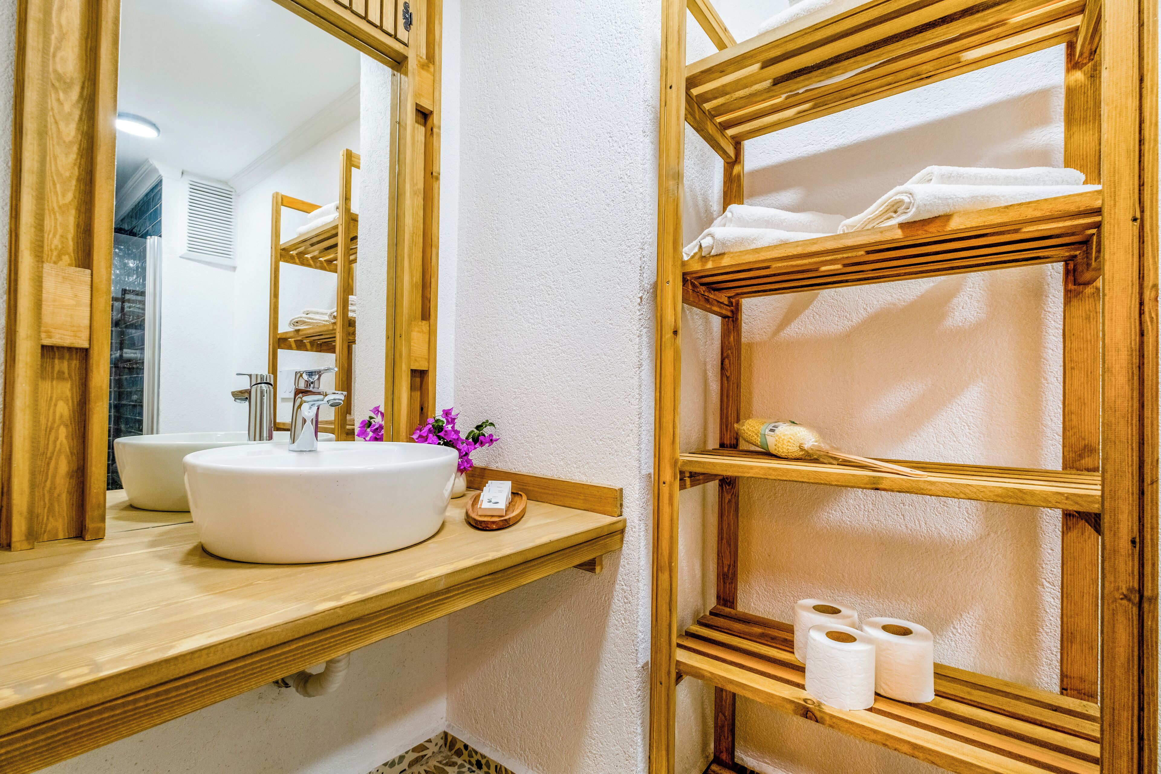 A bathroom with a wooden vanity, a white sink, and a mirror. There are towels and toiletries on the shelves.