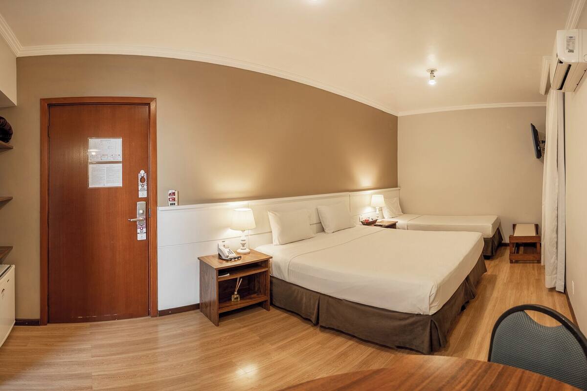 Quarto de hotel com porta de madeira, duas camas, mesa de cabeceira com abajur, uma mesinha e uma cadeira.