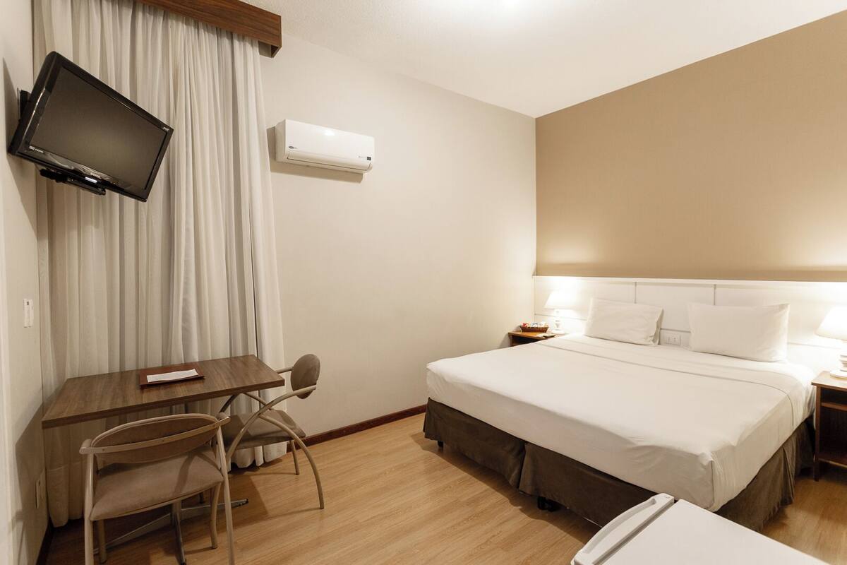 Quarto de hotel com cama, mesas de cabeceira, escrivaninha, cadeira, televisão e ar condicionado.