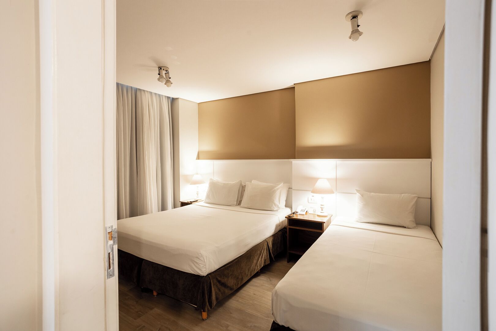 Quarto de hotel com duas camas, uma mesa de cabeceira com abajur e luminária no teto.