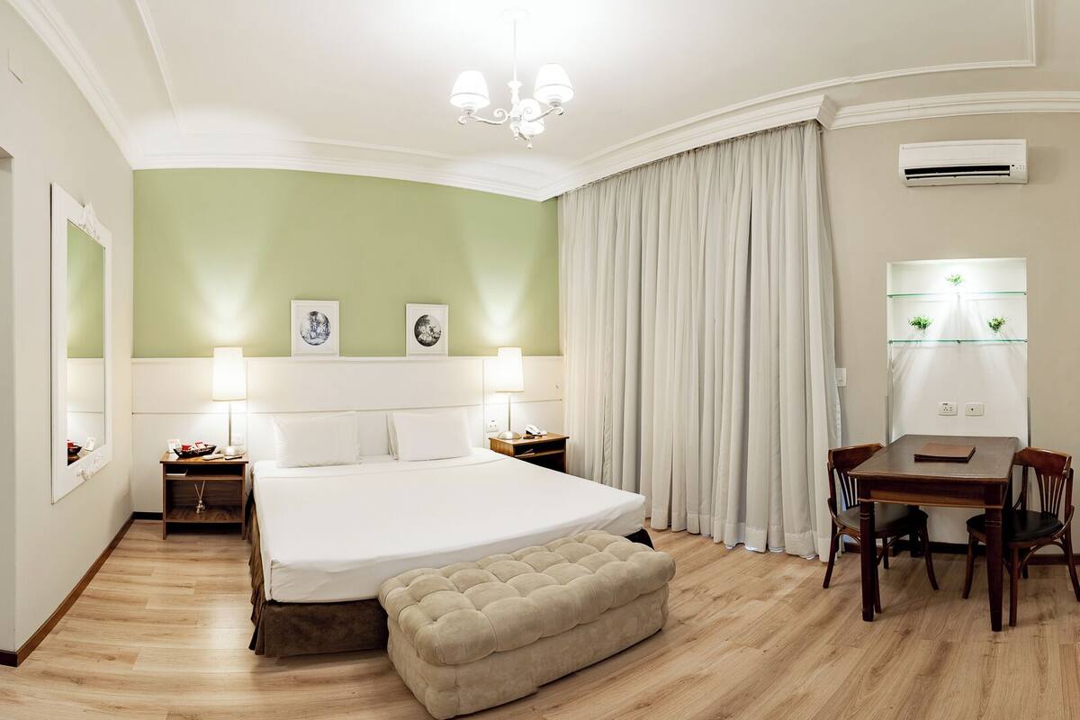 Quarto de hotel com uma cama grande, uma escrivaninha com duas cadeiras, uma mesa de jantar, uma janela ampla com vista e um ar condicionado.