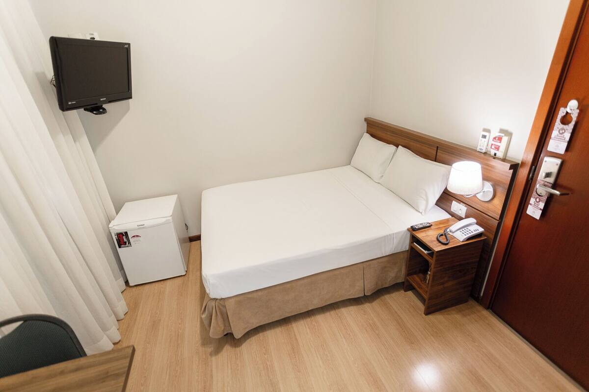 Quarto de hotel com cama, televisão fixada na parede, mesa de cabeceira com abajur, telefone e minibar.