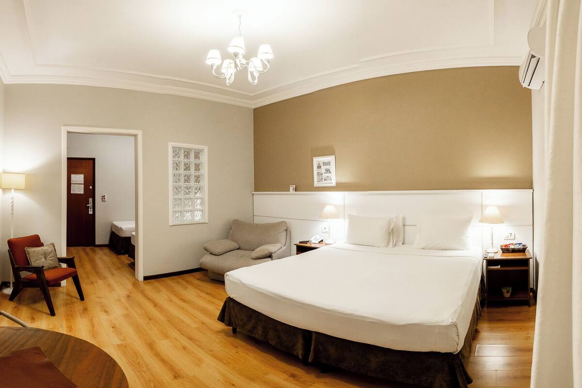 Quarto de hotel com uma cama grande, uma escrivaninha, uma cadeira, uma televisão e um candelabro.