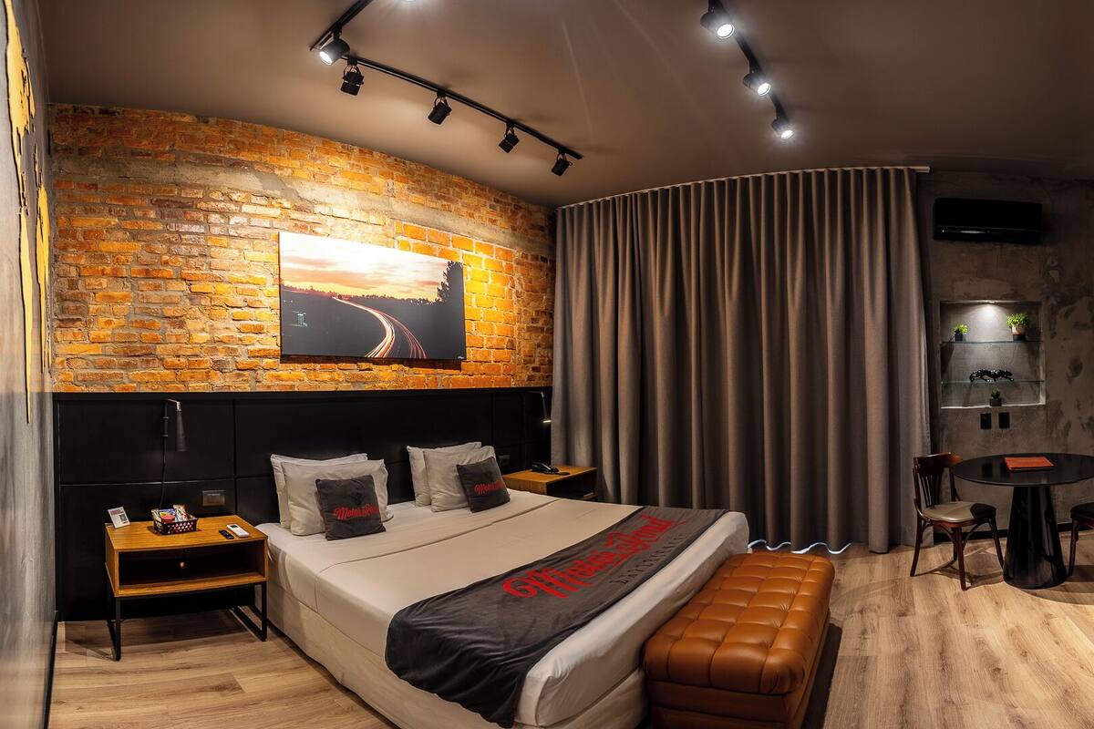 Quarto de hotel moderno com parede de tijolos, uma cama grande, uma escrivaninha com duas cadeiras, uma planta em vaso e uma TV fixada na parede.