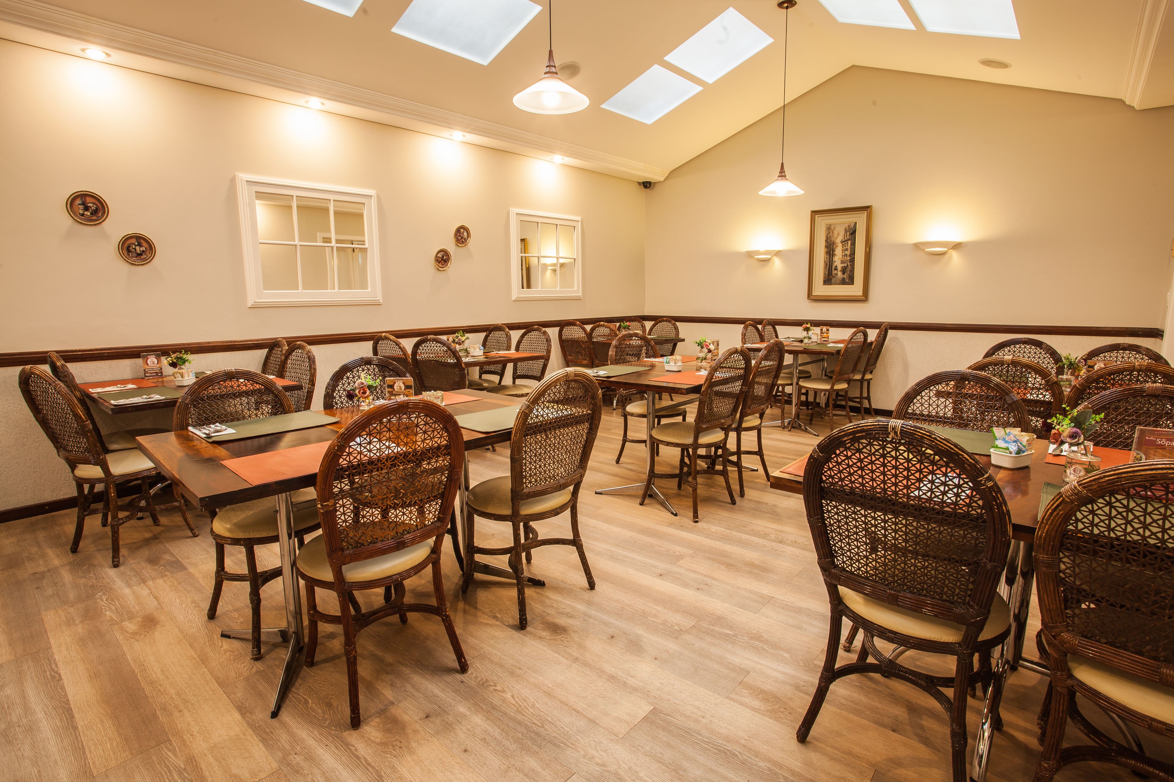 A dining area with multiple tables and wicker chairs, featuring skylights and wall-mounted lights.