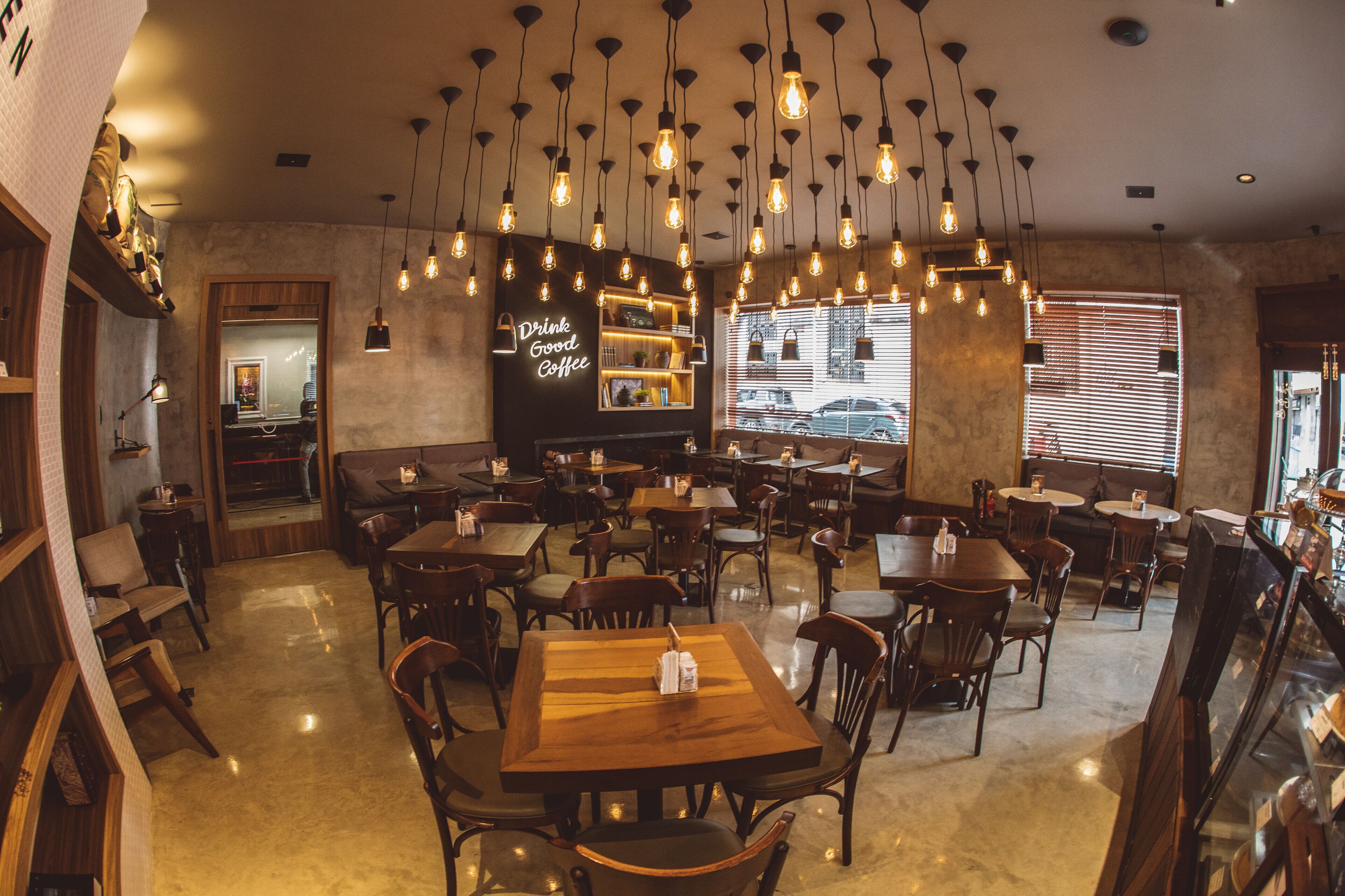 Un café acogedor con mesas y sillas de madera, lámparas colgantes y un letrero que dice 'Drink Good Coffee'.