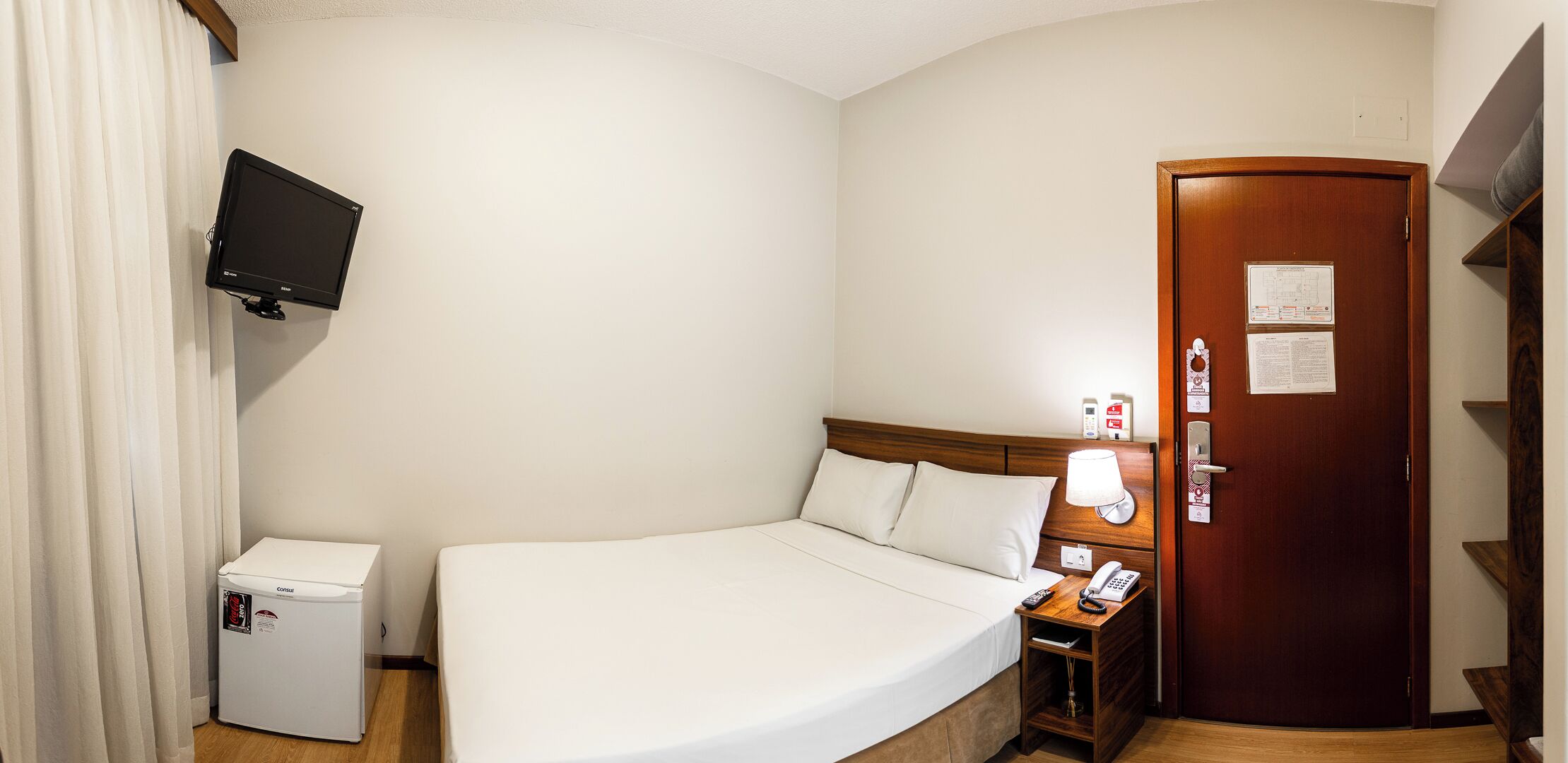 Una habitación de hotel con una cama, una mesita de noche con lámpara, un teléfono, un refrigerador pequeño y una puerta de madera con un aviso.