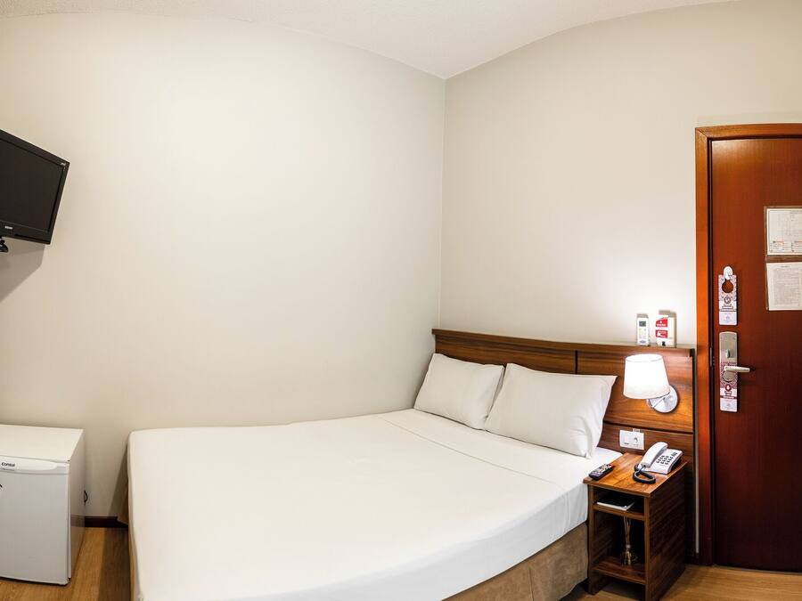Una habitación de hotel con una cama, una mesita de noche con lámpara, un teléfono, un refrigerador pequeño y una puerta de madera con un aviso.