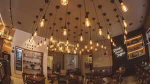 Interior de una cafetería con focos colgantes, mobiliario de madera y un refrigerador empotrado en la pared.