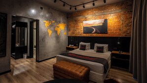 Un dormitorio moderno con pared de ladrillo, un mural de mapa mundial, una cama grande con cabecera negra y un cuadro de una carretera al atardecer.