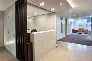 Zona de recepción con mostrador de mármol, puerta de madera y pasillo alfombrado que conduce a zonas de estar.