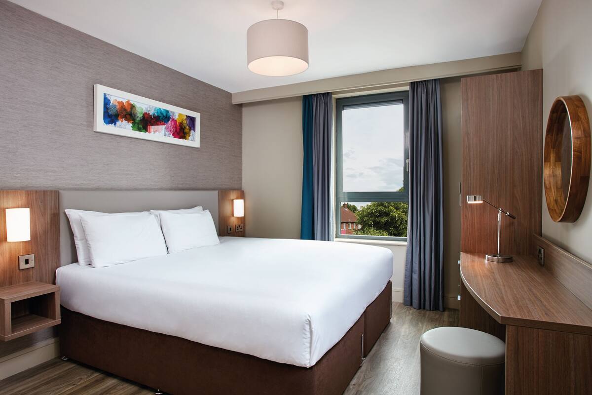 A modern hotel room with a large bed, bedside lamps, a desk, and a window with curtains.