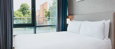 A hotel room with a large bed, a view of buildings and trees through a window, and a colorful artwork on the wall.