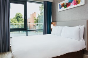 A hotel room with a large bed, a view of buildings and trees through a window, and a colorful artwork on the wall. - Cordia Serviced Apartments (Belfast)