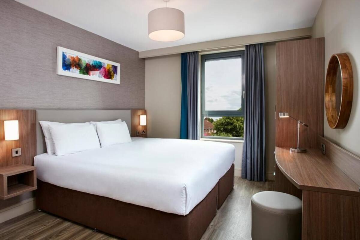 A hotel room with a large bed, bedside lamps, a desk with a lamp, a trash bin, and a large window with curtains.