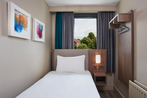 Una habitación de hotel con cama, mesita de noche, lámpara de pared, ventana con cortinas y dos cuadros enmarcados.