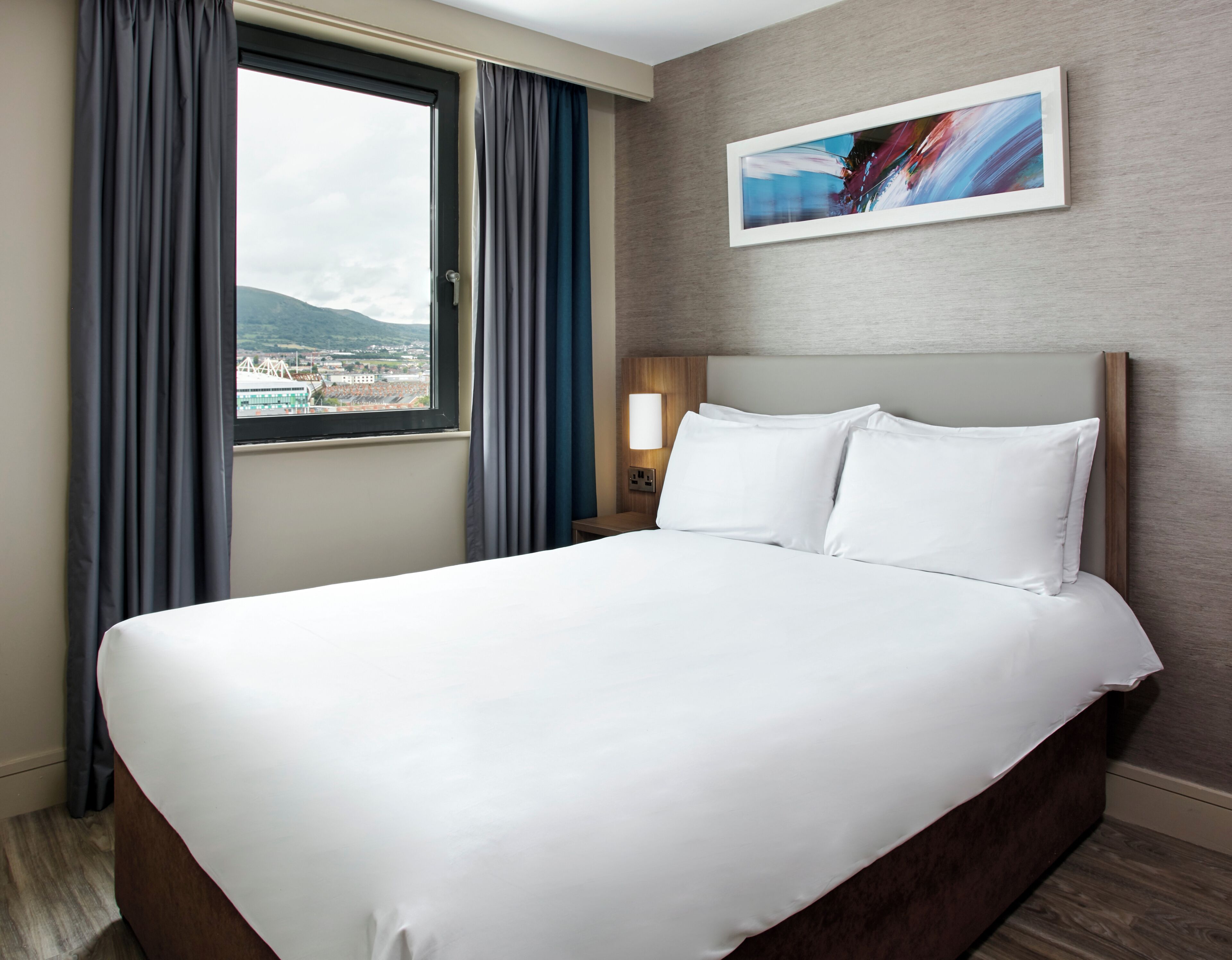 A hotel room with a large bed, a window with curtains, a nightstand with a lamp, and a painting on the wall.