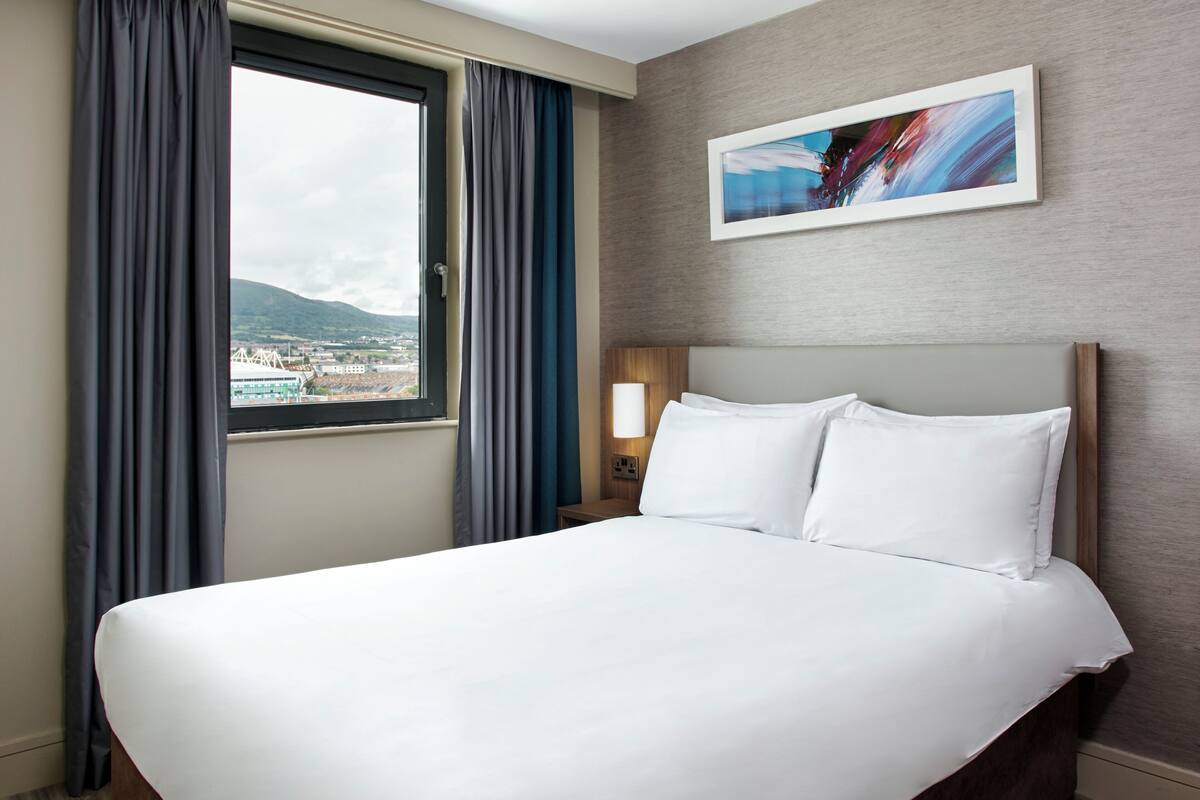 A hotel room with a large bed, a window with curtains, a nightstand with a lamp, and a painting on the wall.