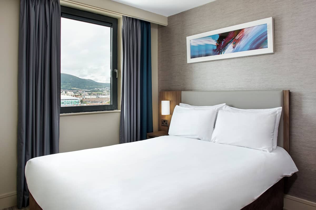 Un cuarto de hotel con una cama grande, una ventana con cortinas, una mesita de noche con lámpara y un cuadro en la pared.