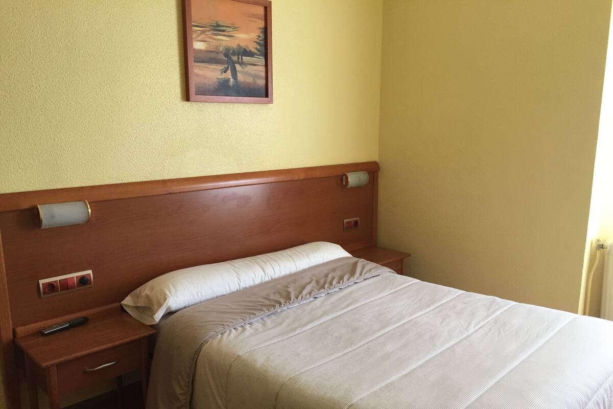 A hotel room with a wooden headboard, a single bed with a beige bedspread, a bedside table with a remote control, and a wall-mounted light switch.
