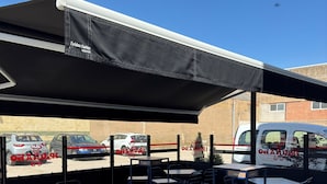 An outdoor seating area with tables and chairs, a covered section, and a view of parked cars in the background. - The Hostal Iruñako (Burgos)