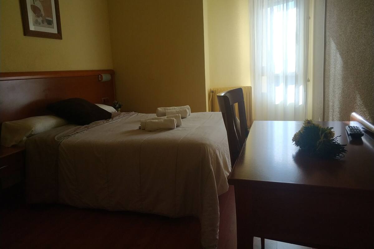 A hotel room with a bed, a wooden headboard, a table with a remote control, and a window with curtains.
