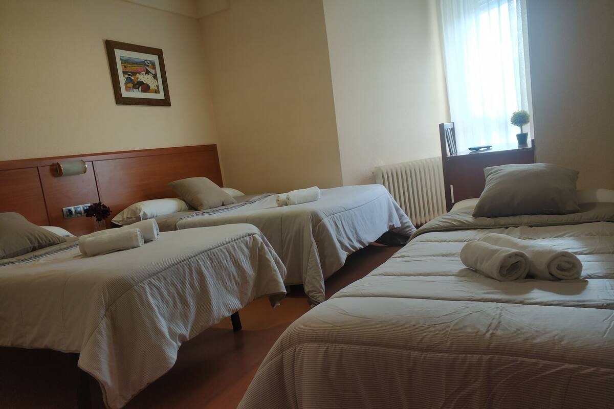 A hotel room with two single beds and a double bed, each with neatly made linens and pillows. There is a framed picture on the wall, a radiator, and a window with curtains.