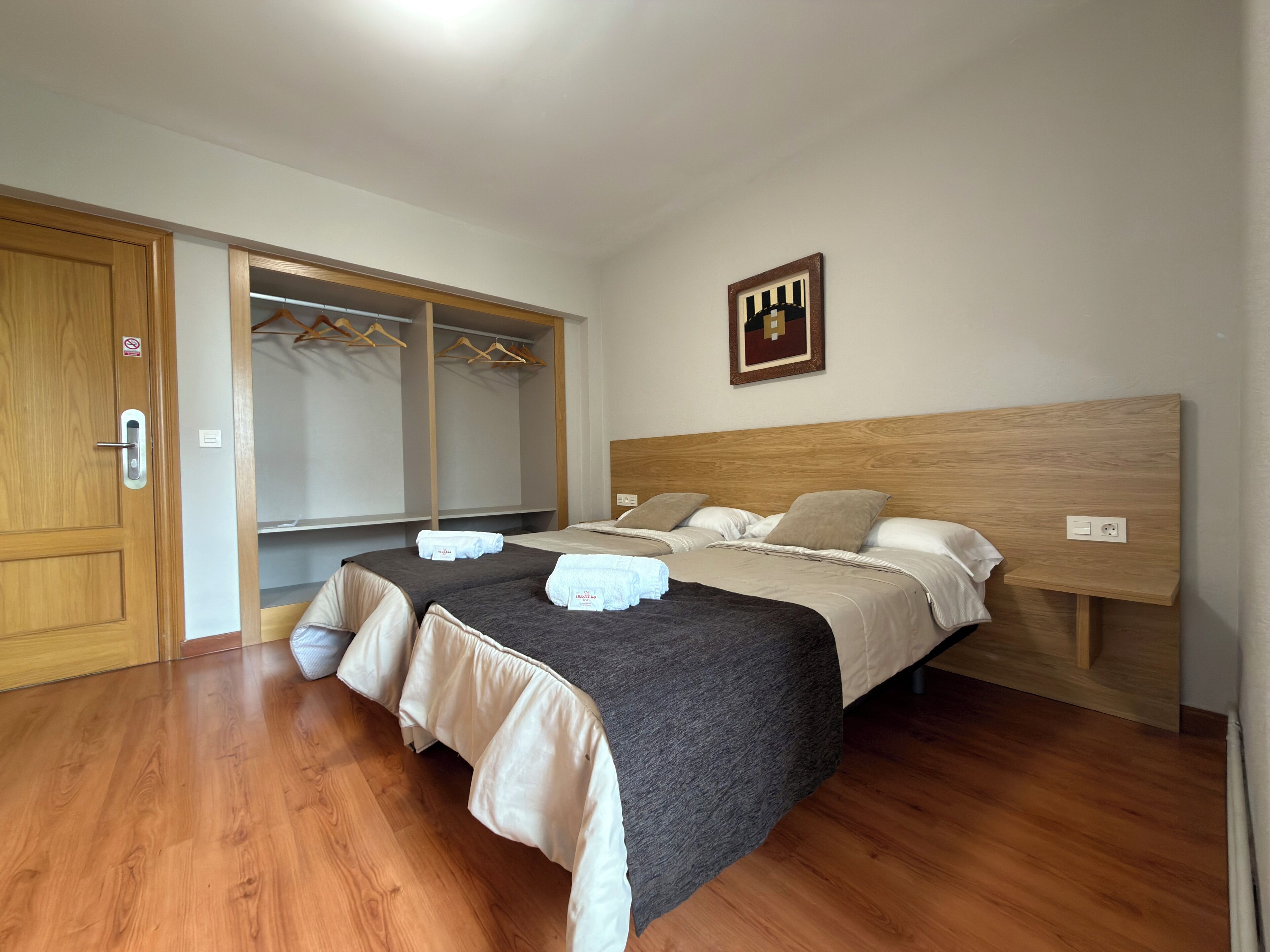 Une chambre d’hôtel avec deux lits, une tête de lit en bois, une armoire avec espace pour suspendre les vêtements et une œuvre d’art accrochée au mur.