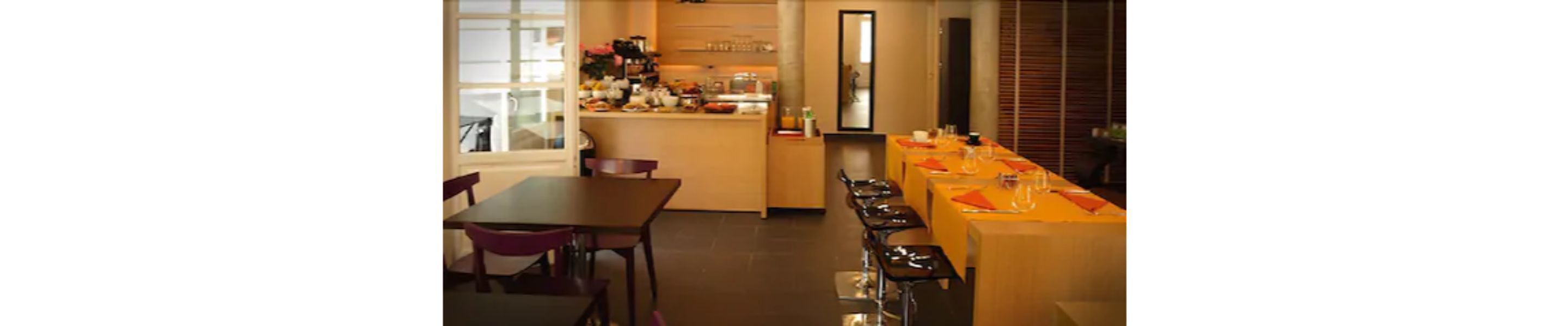 A hotel dining area with a long table set for a meal, a kitchen area with a buffet, and a seating area with chairs.