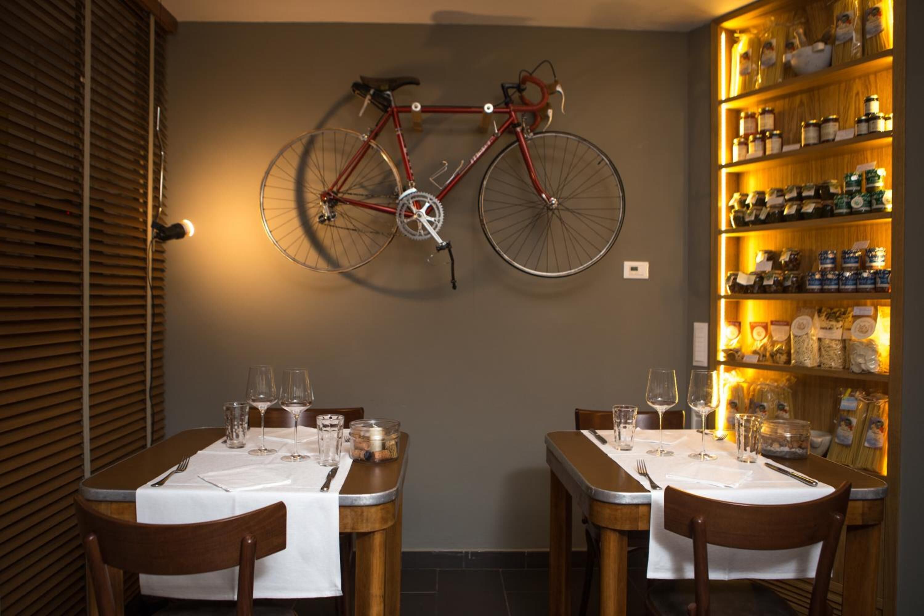 Un coin repas avec deux tables préparées pour deux personnes, un vélo rouge accroché au mur et, à droite, des étagères remplies de pots et de bouteilles.