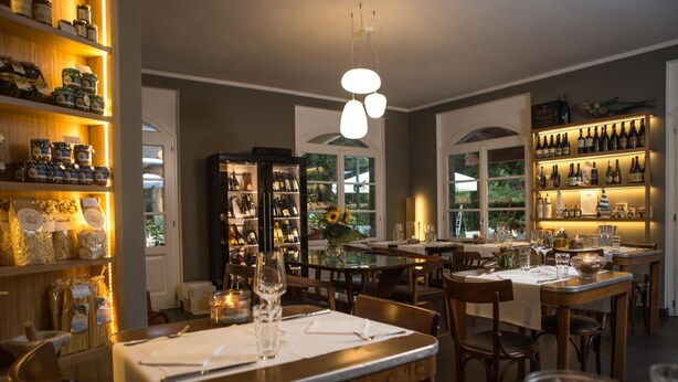 Un restaurante con varias mesas, estantes llenos de frascos y botellas, y vistas al exterior con vegetación.
