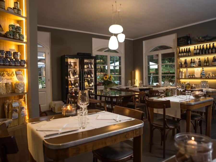 A restaurant with multiple tables, shelves with jars and bottles, and a view of greenery outside.