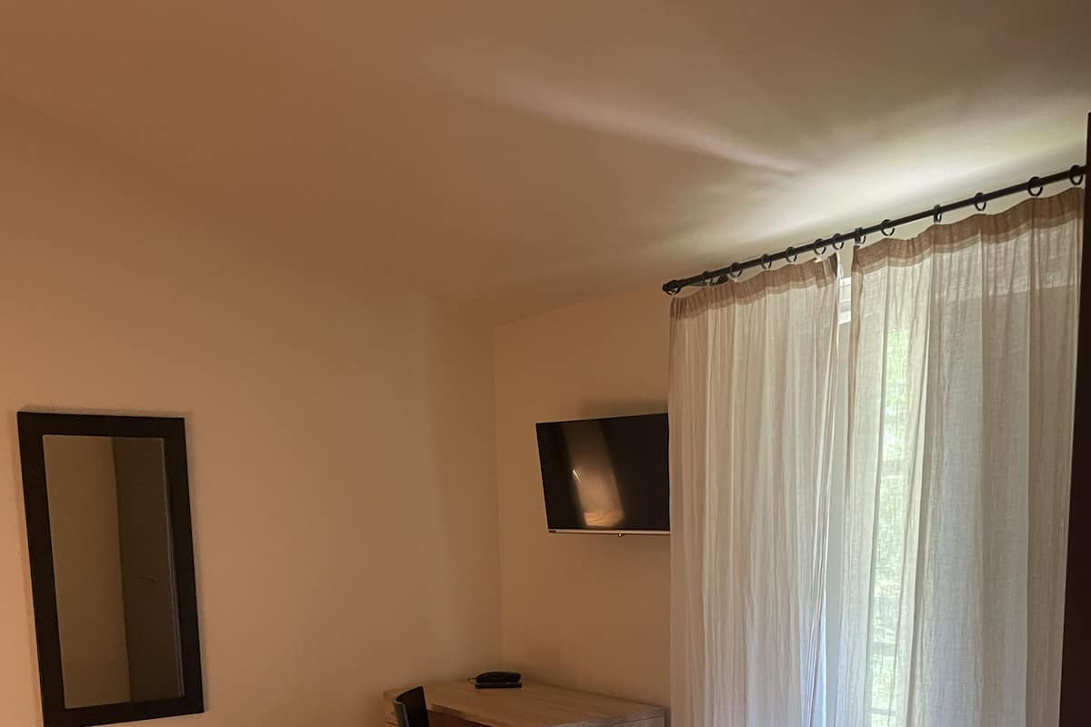 A room with a bed, a desk, a chair, a TV, and a window with sheer curtains.