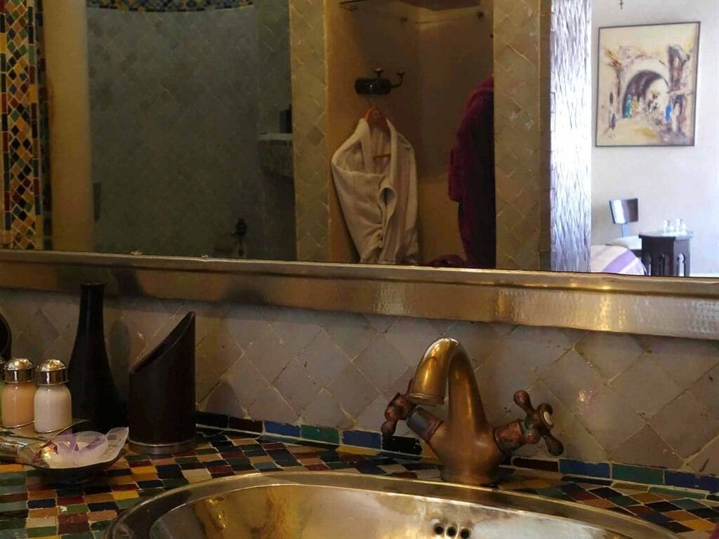 A bathroom with a tiled backsplash, a sink with a gold faucet, and a mirror reflecting a towel and a painting on the wall.