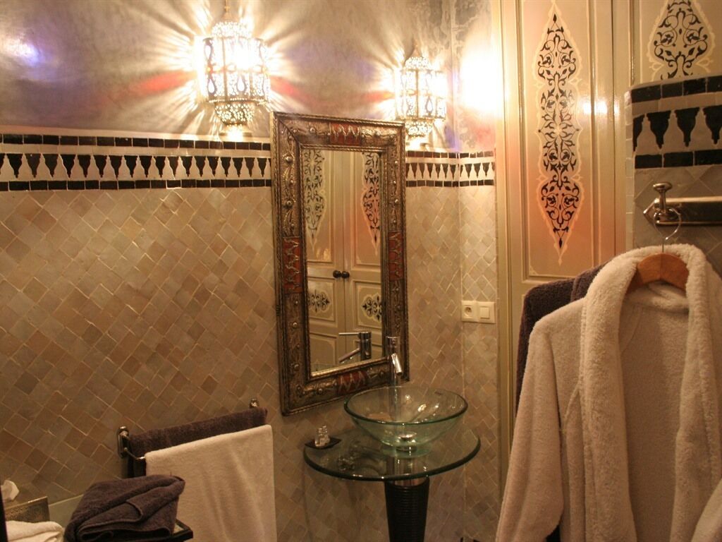 A bathroom with a glass sink, two wall-mounted lights, a decorative mirror, and patterned tile walls.