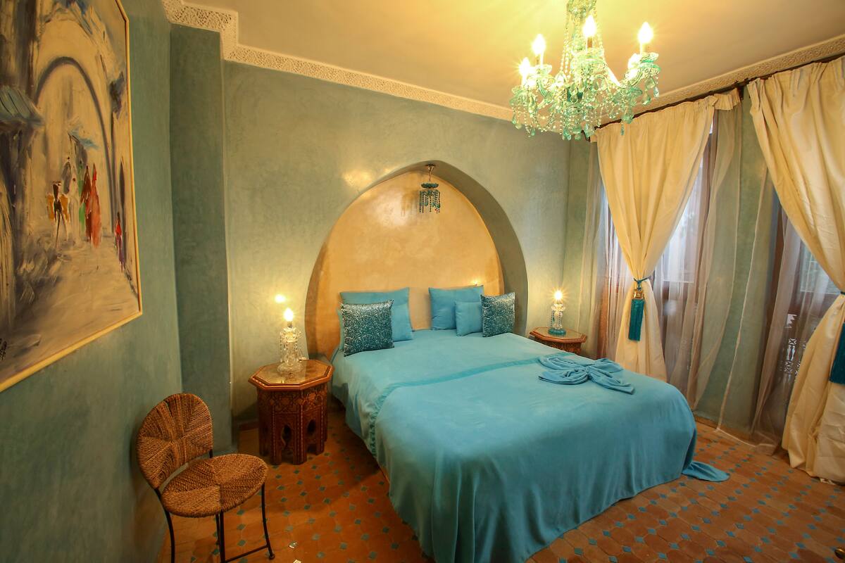 A bedroom with a large bed, a chair, a painting on the wall, and a chandelier.