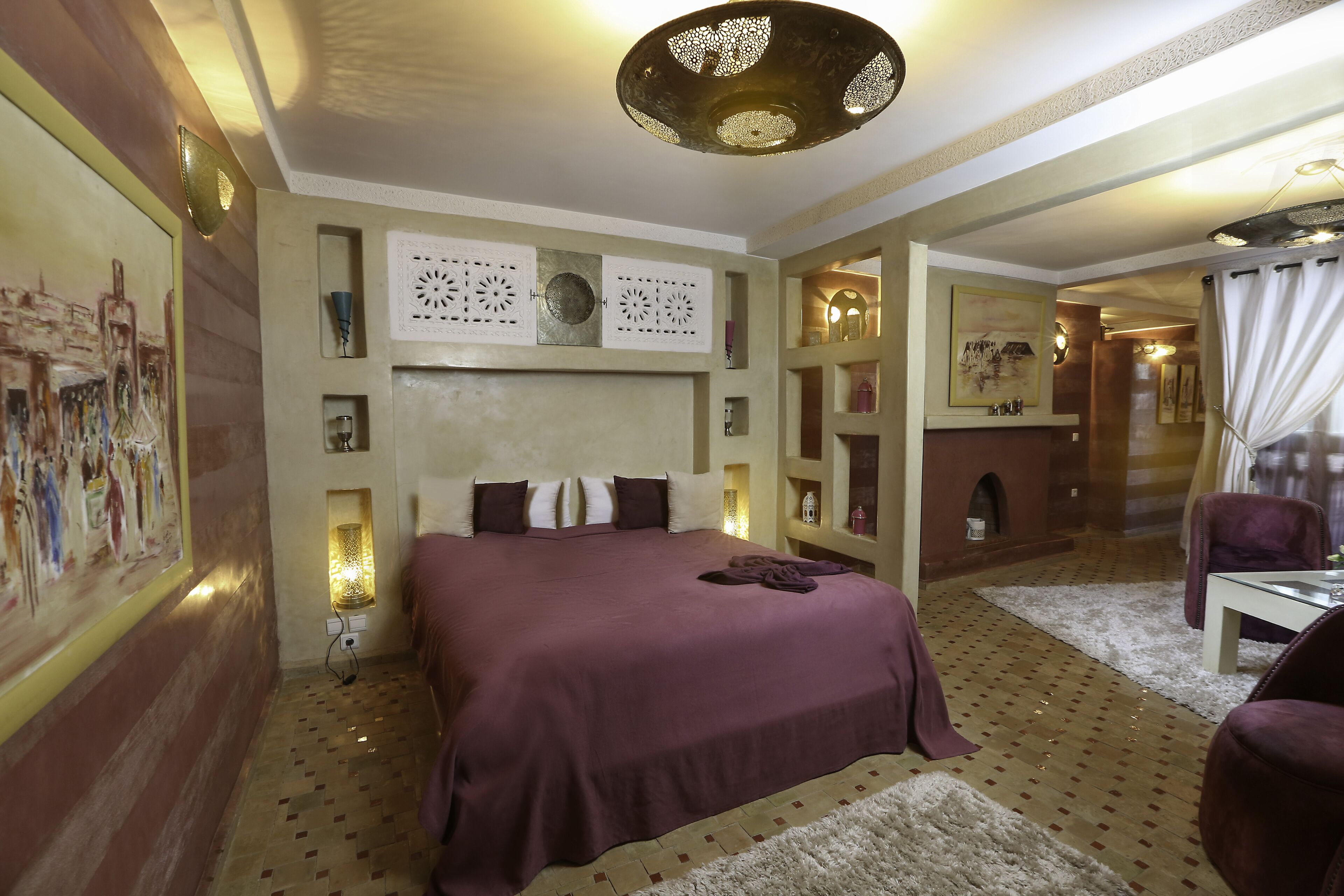 Un dormitorio con una cama grande, una mesita pequeña y una silla. Hay decoraciones en la pared y una lámpara en el techo.