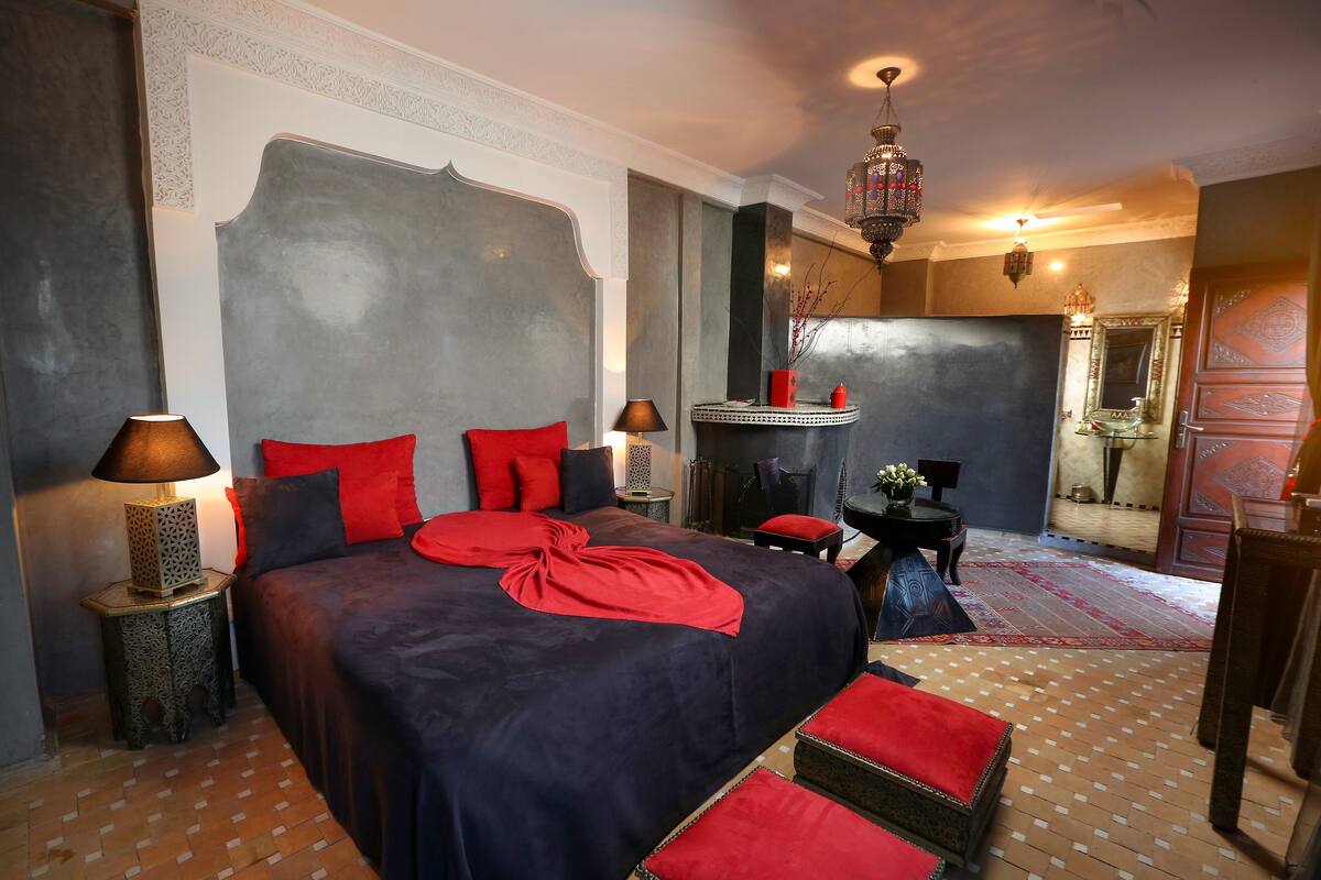 A bedroom with a large bed, red pillows, a red throw, a nightstand with a lamp, and a wooden floor.
