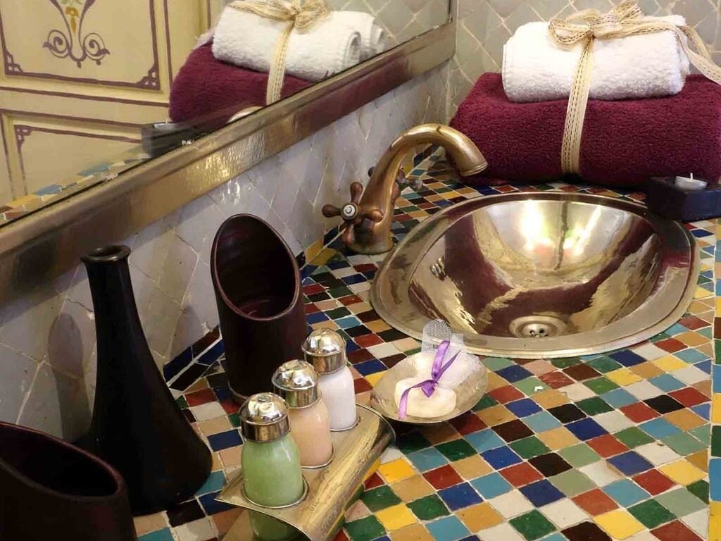 Un baño con un piso de mosaicos de colores vivos, un lavamanos grande con grifo dorado y toallas ordenadas en un estante.