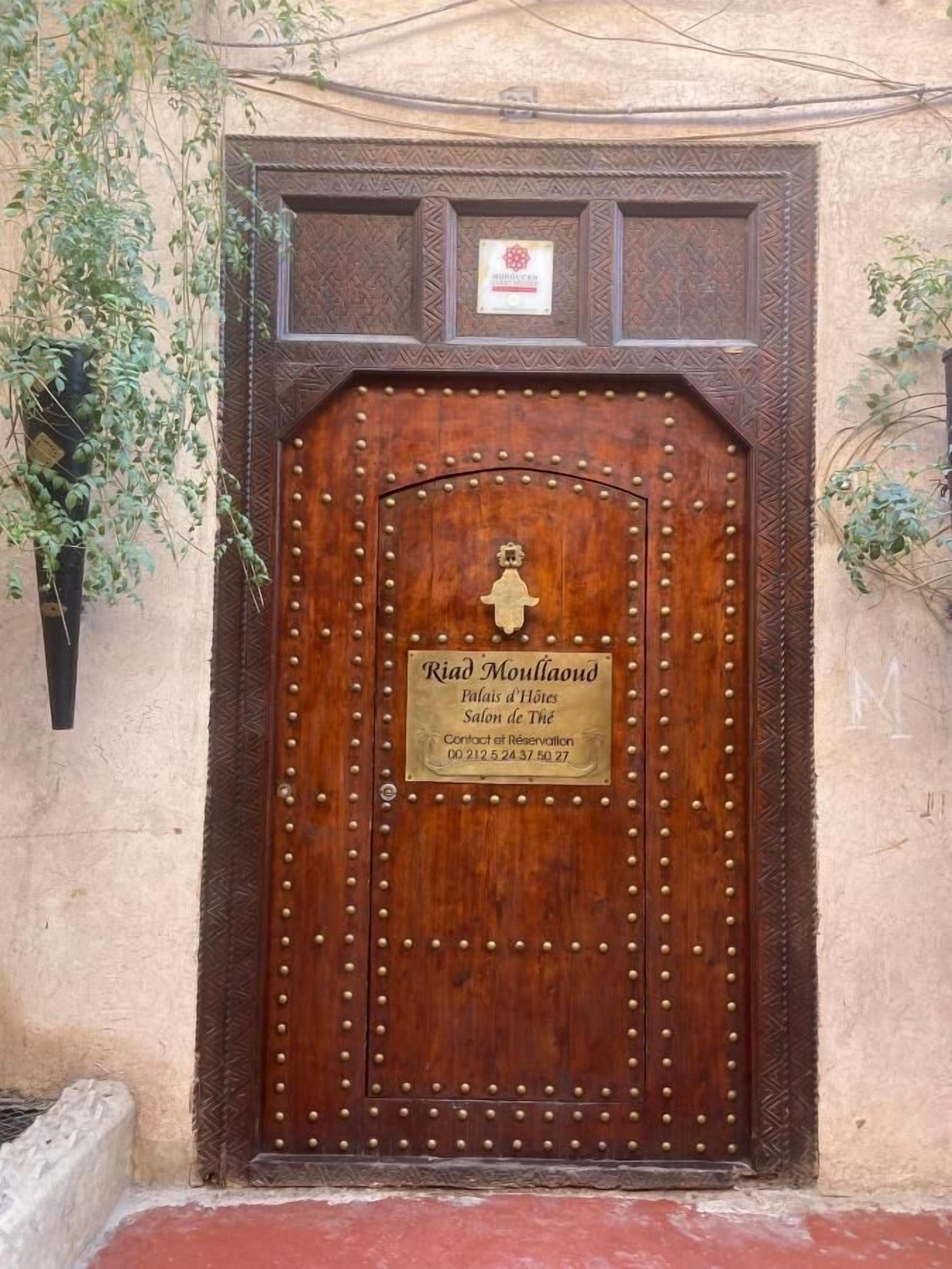 Eine Holztür mit einer Messingklopfer und einer Schilder mit der Aufschrift "Riad Moullaoud" und Kontaktinformationen.