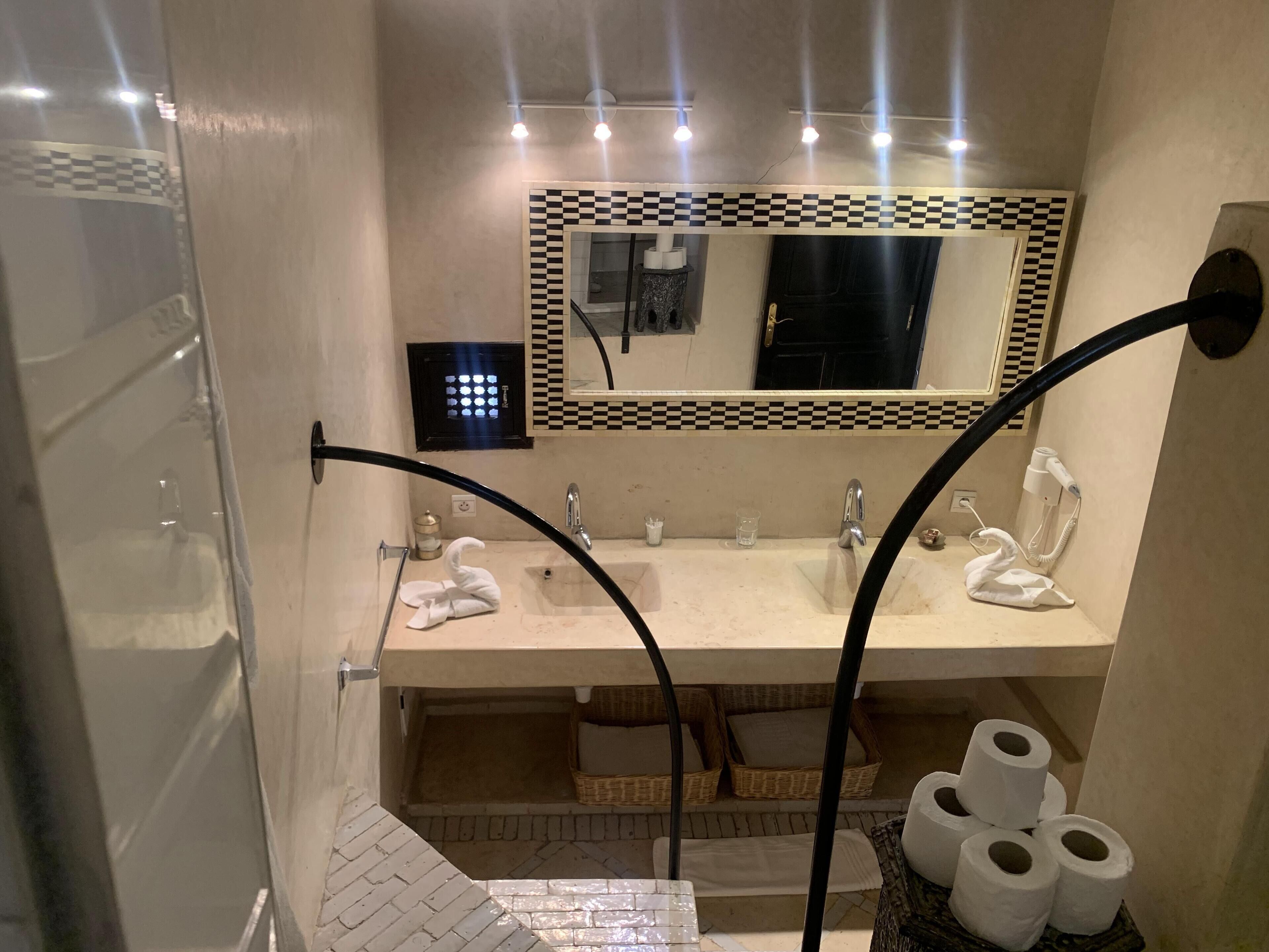A bathroom with a large mirror, two sinks, a towel rack, and a basket of toilet paper.