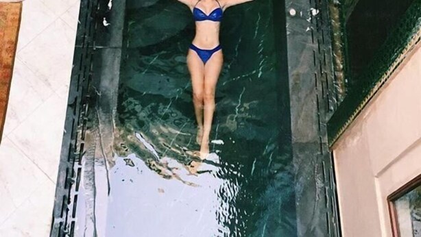 Una mujer con traje de baño azul parada en una fuente de agua con un muro de piedra y una planta encima.