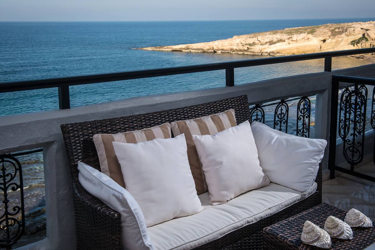 A wicker sofa with white cushions on a balcony overlooking a rocky coastline.