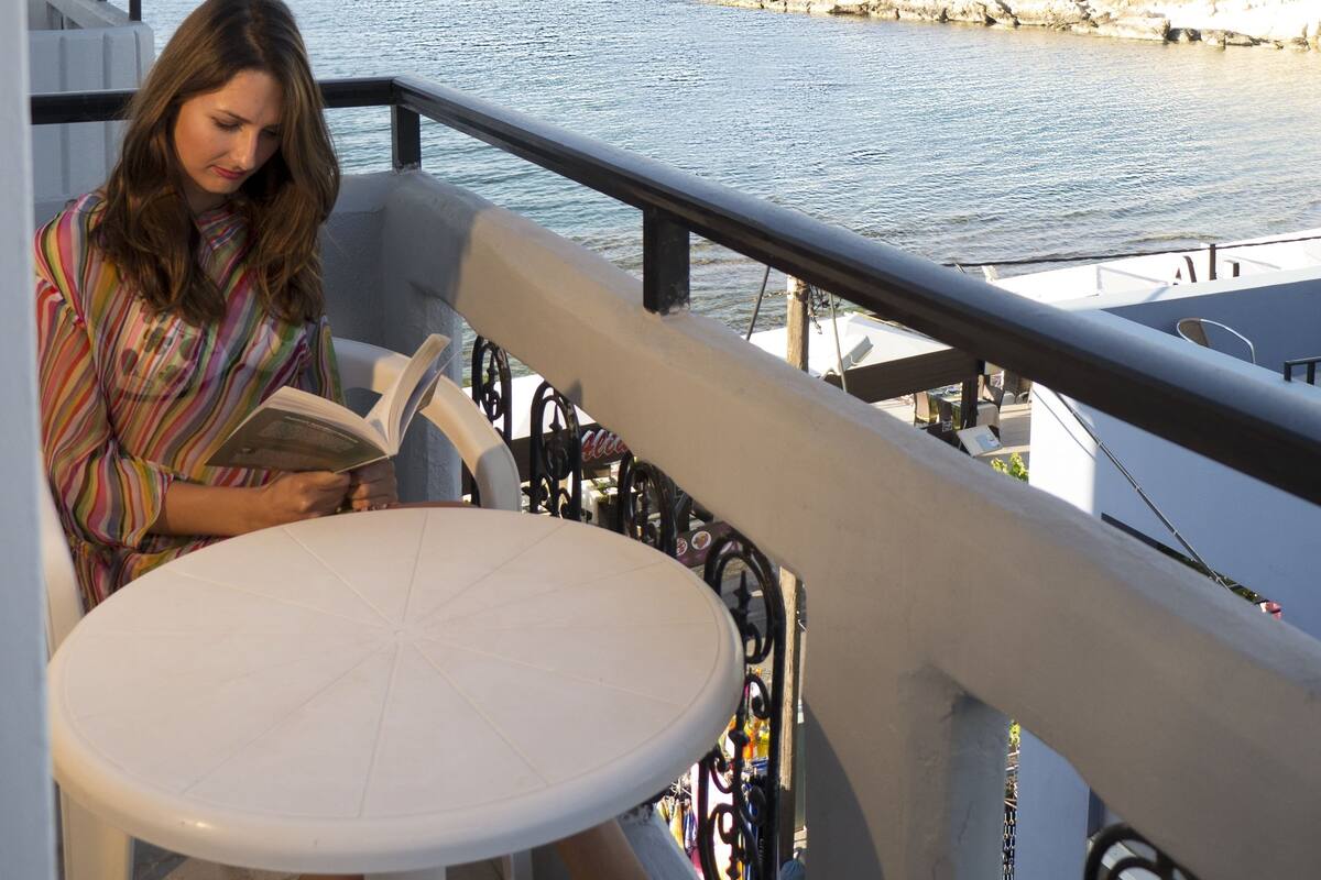 A balcony with a white table and a person reading a book. The balcony overlooks a rocky coastline.