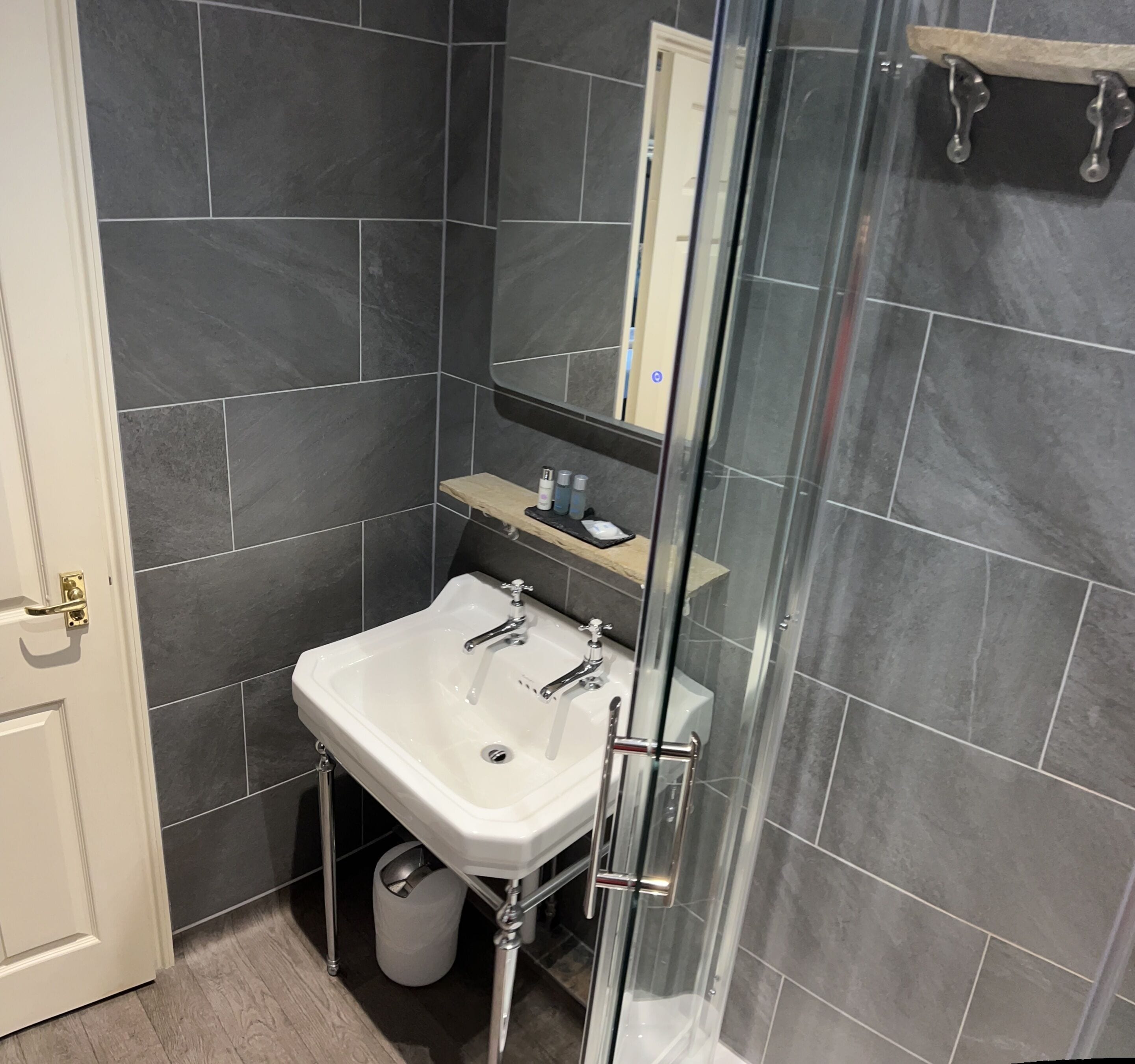 A bathroom with a white sink, dark tiles, a glass shower enclosure, and a wooden shelf with toiletries.