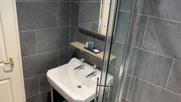 A bathroom with a white sink, dark tiles, a glass shower enclosure, and a wooden shelf with toiletries.