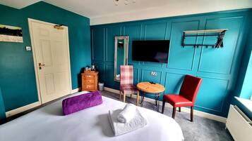 A bedroom with teal walls, a bed with white linens, a red chair, a small round table, a wooden dresser, and a TV mounted on the wall.