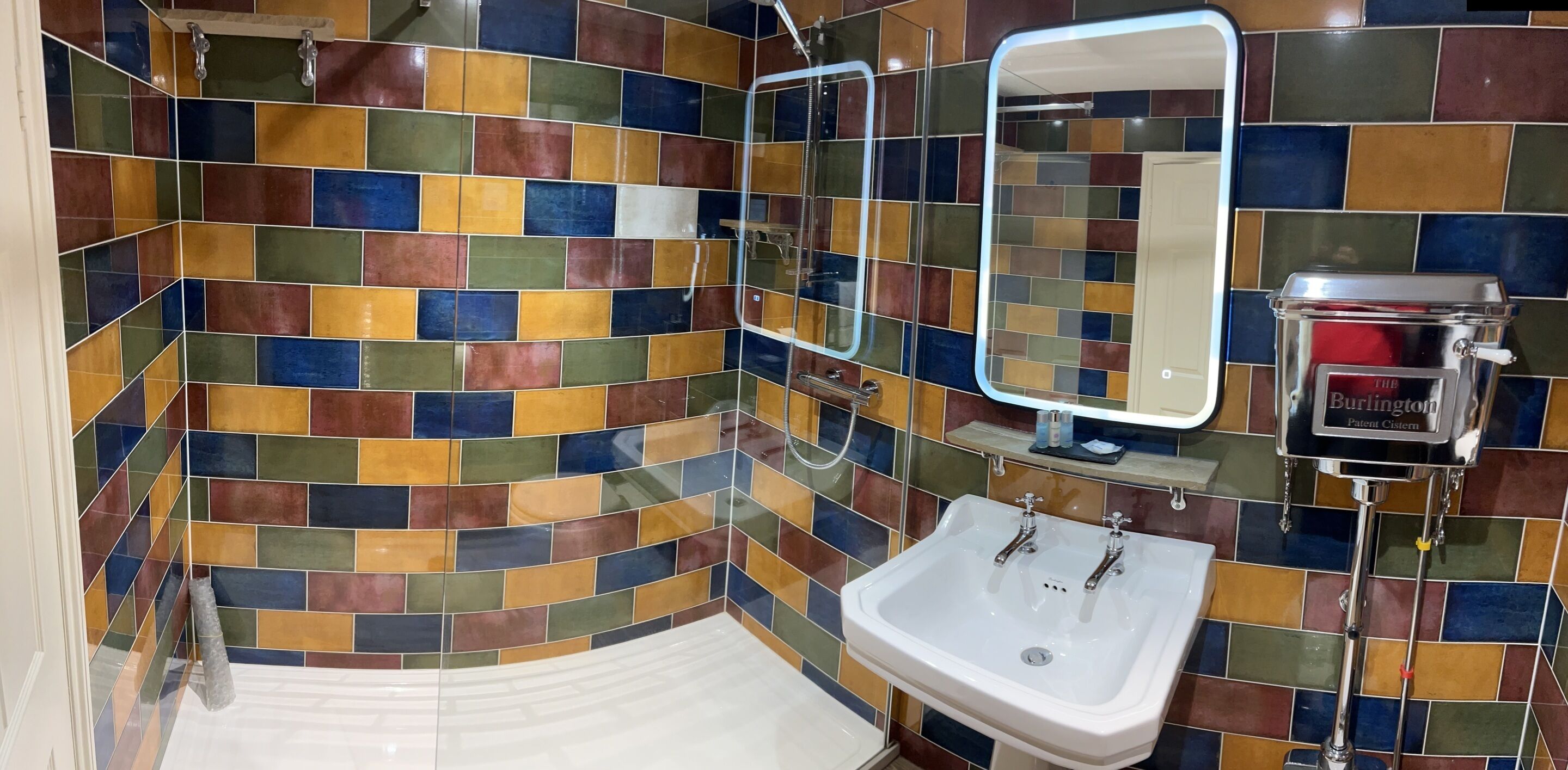 Un baño con mosaicos de colores, un lavamanos blanco, una cabina de ducha con paredes de vidrio y un espejo con dispensador de jabón rojo.