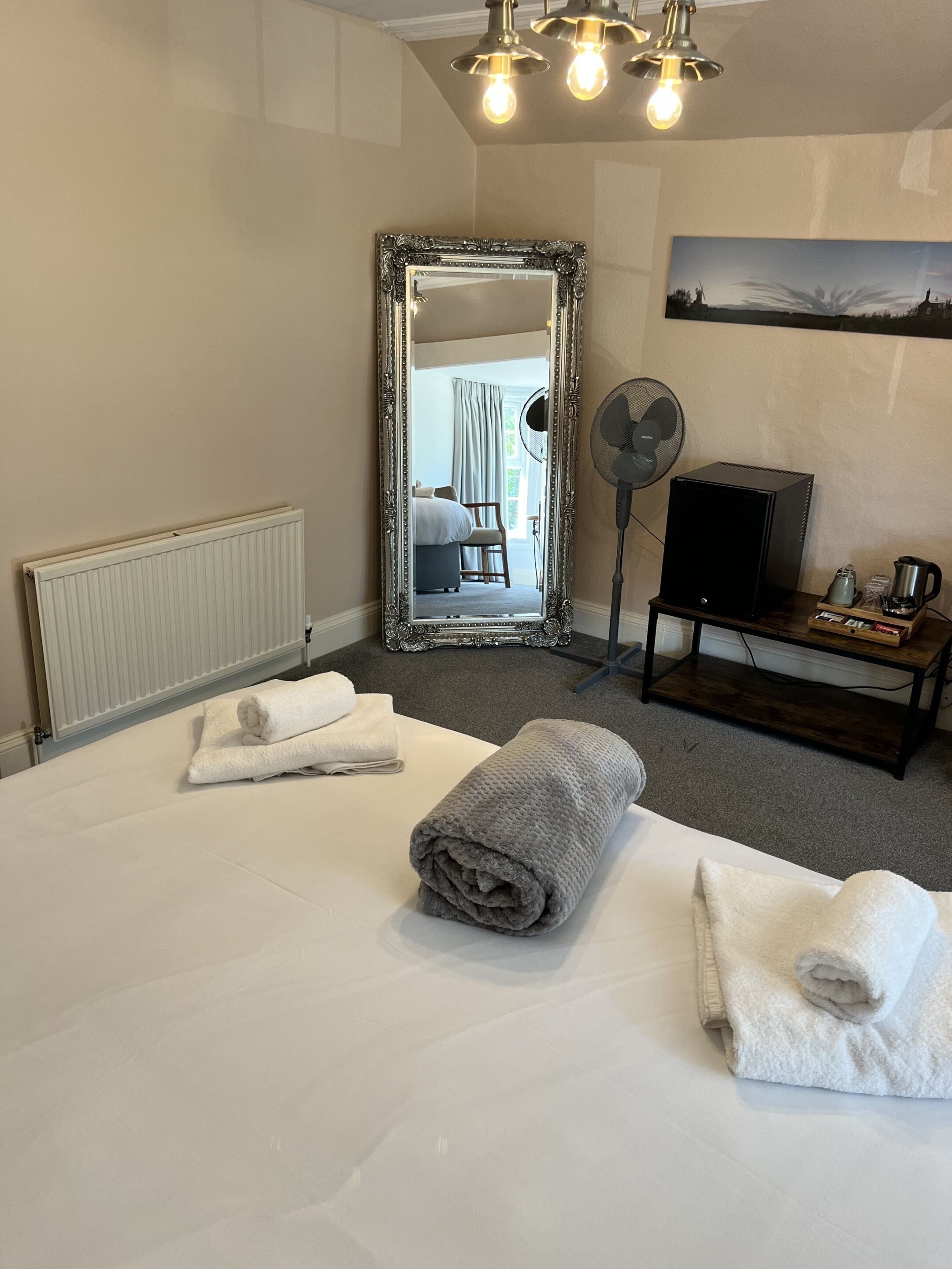 A hotel room with a bed, two rolled towels, a mirror, a fan, a coffee maker, and a painting on the wall.