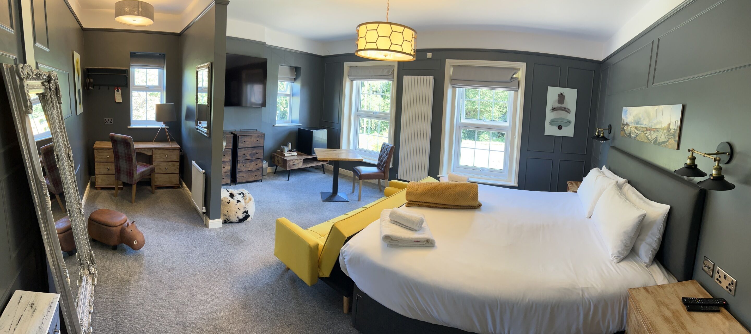 A hotel room with a large bed, a desk, a chair, a sofa, and a small table. There are windows with curtains, a mirror, and artwork on the walls.